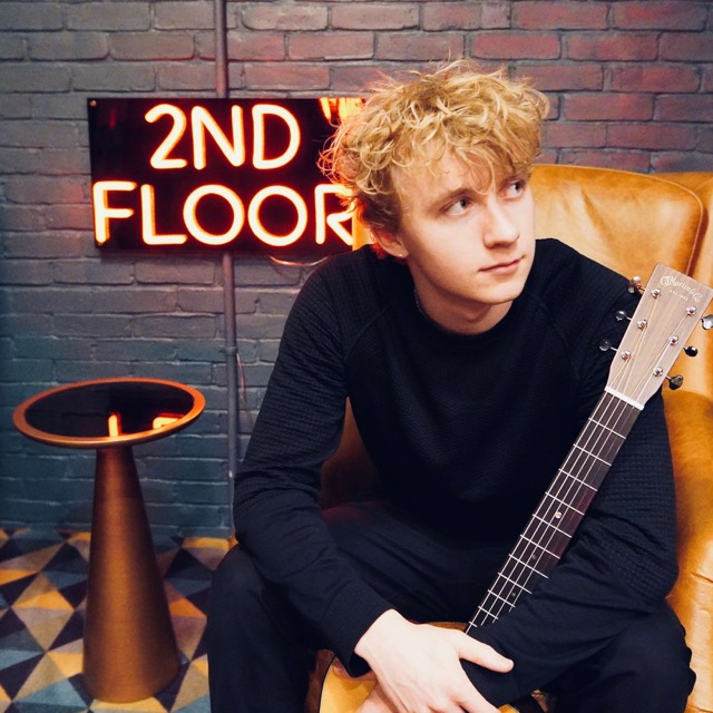 Takoda Dionne seated with an acoustic guitar under a neon sign