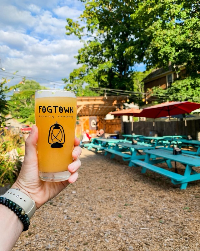 A hand raising a Fogtown glass of hazy beer over the garden’s turquoise picnic tables