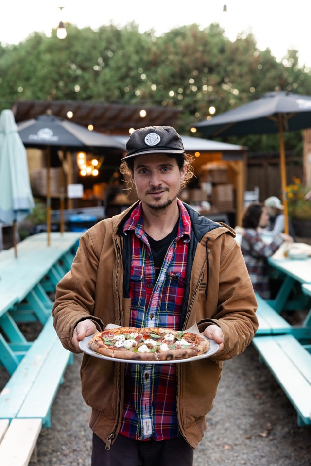 A cook in a flannel shirt and cap holding up a wood-fired pizza in the beer garden