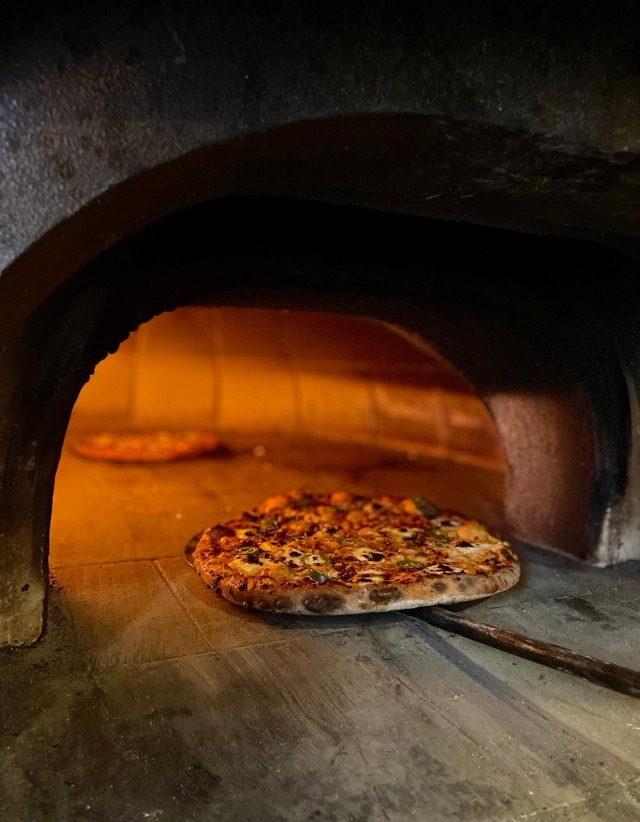 A pizza baking on the floor of the glowing wood-fired oven