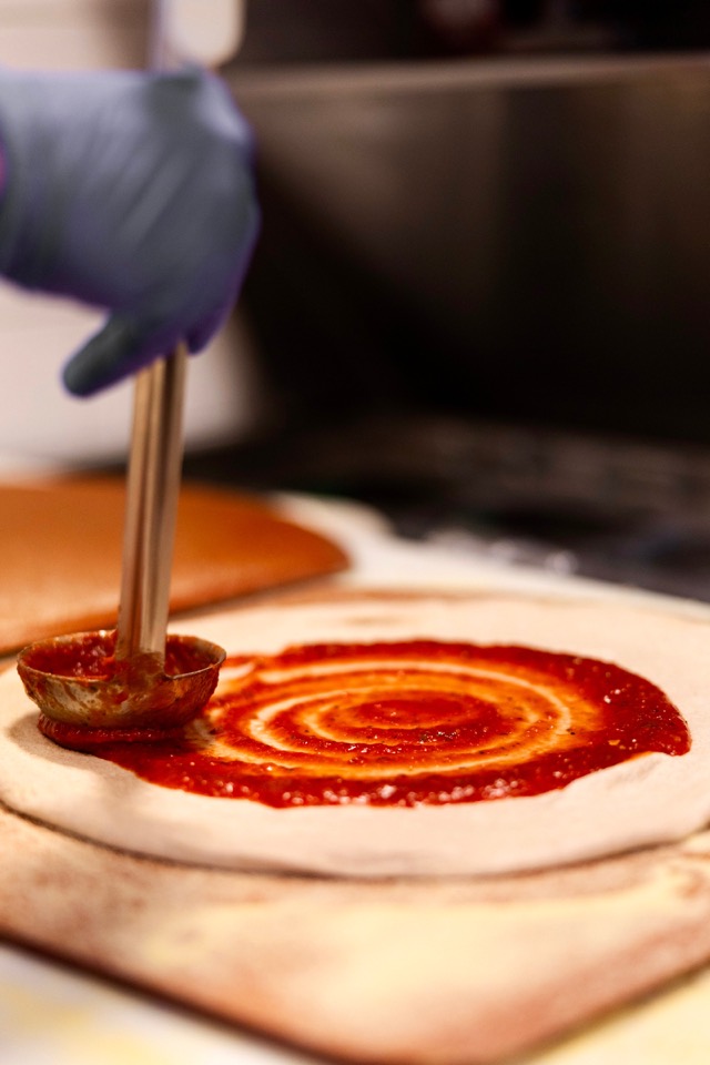 A ladle spiraling red sauce onto stretched pizza dough